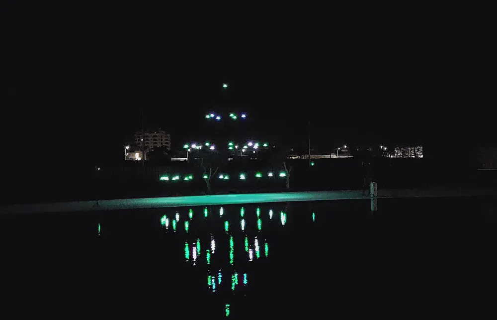 Espetáculo noturno de drones com luzes verdes refletidas na água, formando padrões geométricos sincronizados.